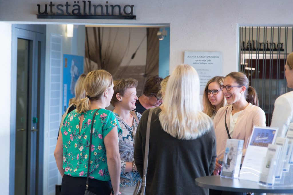 Joukko naisia ​​seisoo ja juttelee näyttelykyltin lähellä hyvin valaistussa sisätiloissa, luultavasti seminaarikonferenssi- ja juhlasalissa, etualalla esitteitä.