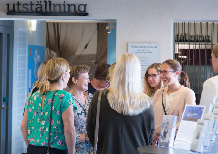 En grupp kvinnor står och pratar nära en utställningsskylt i en väl upplyst inomhusmiljö, troligen på en seminariekonferens och ett juhlalle, med broschyrer synliga i förgrunden.