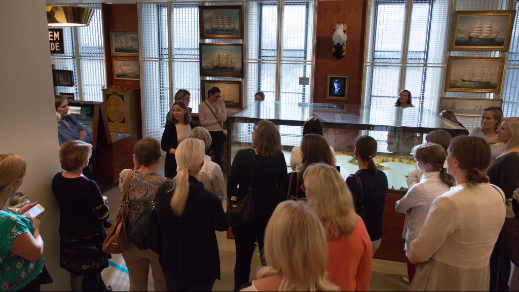 A group of people listen to a guide in a museum room with ship paintings, exhibits and displays.