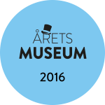 Blått runt märke som visar ÅRETS MUSEUM 2016 i svart text, med en illustration av en hög hatt ovanför bokstaven A i ÅRETS, vilket symboliserar ett prestigefyllt museipris.