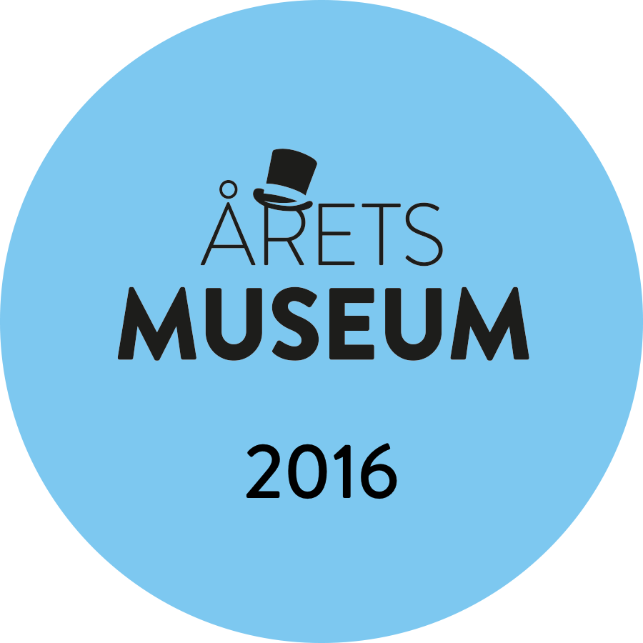 Sininen pyöreä rintanappi, jossa näkyy VUODEN MUSEO 2016 mustalla tekstillä ja silinterikuva A-kirjaimen yläpuolella ARETS-kirjaimessa, joka symboloi arvostettua museopalkintoa.
