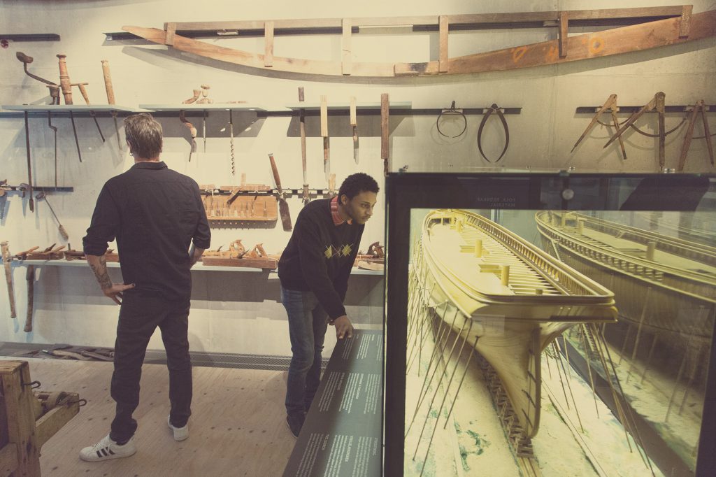 Två personer betraktar en museiutställning som påminner om ett kuriosakabinett, med träbearbetningsverktyg på väggen och en detaljerad modellskeppsmodell i en vitrinskåp.
