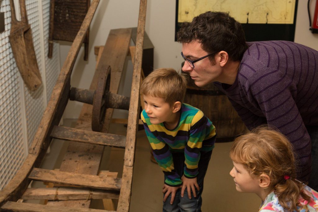 En vuxen och två barn observerar noga ett historiskt träföremål som visas i en museimiljö.