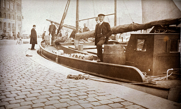 Ett sepiatonat foto av en segelbåt vid kaj med tunnor på däck fångar Segelsjöfartens anda; en man i keps sitter på båten, medan andra står i närheten på kullerstenskajen.