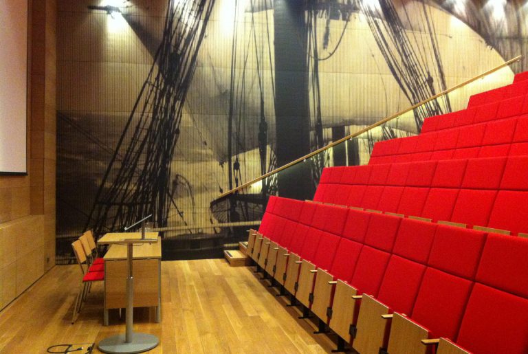 Föreläsningssal med röda sittplatser i flera etager, trägolv och ett podium; bakväggen har en stor väggmålning av ett fartyg med rigg – idealisk seminarium för konferens och fest.