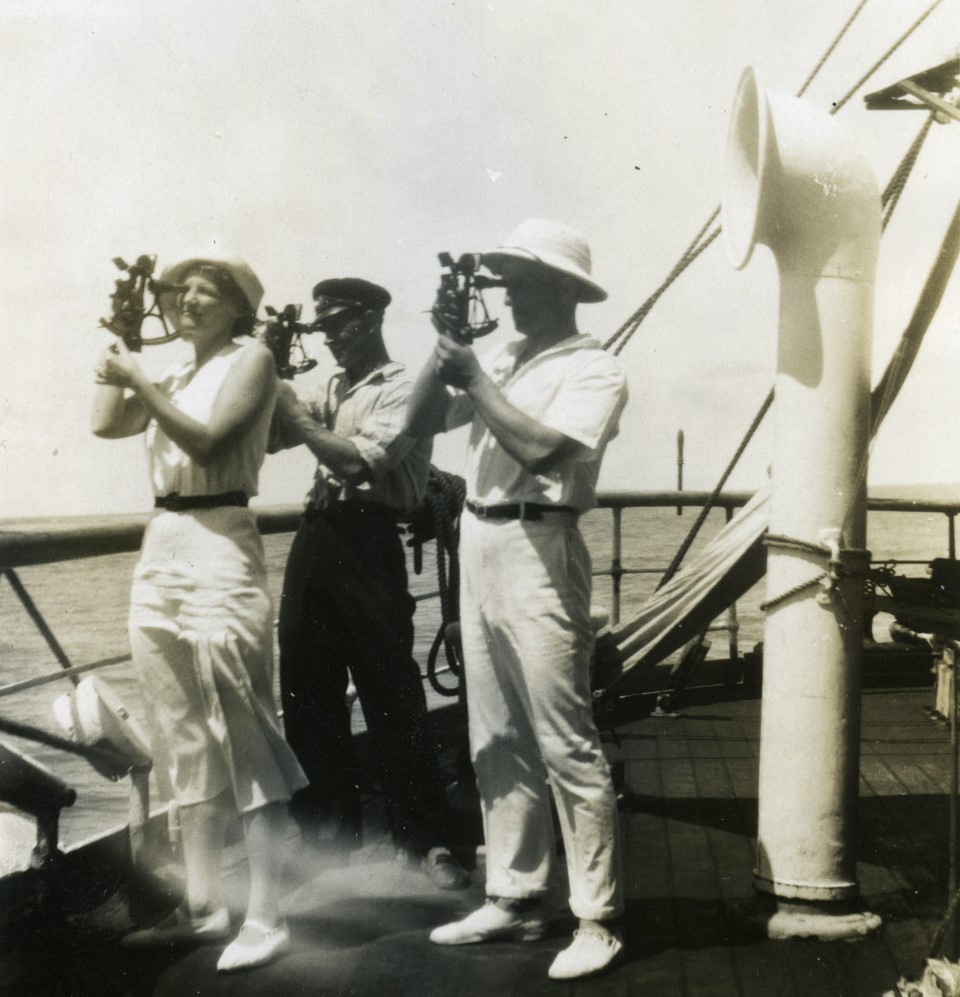 Three people stand on a ship's deck and use sextants for navigation; the sea and part of the ship's structure are visible in the background, highlighting their skillful approach to classical navigational methods.