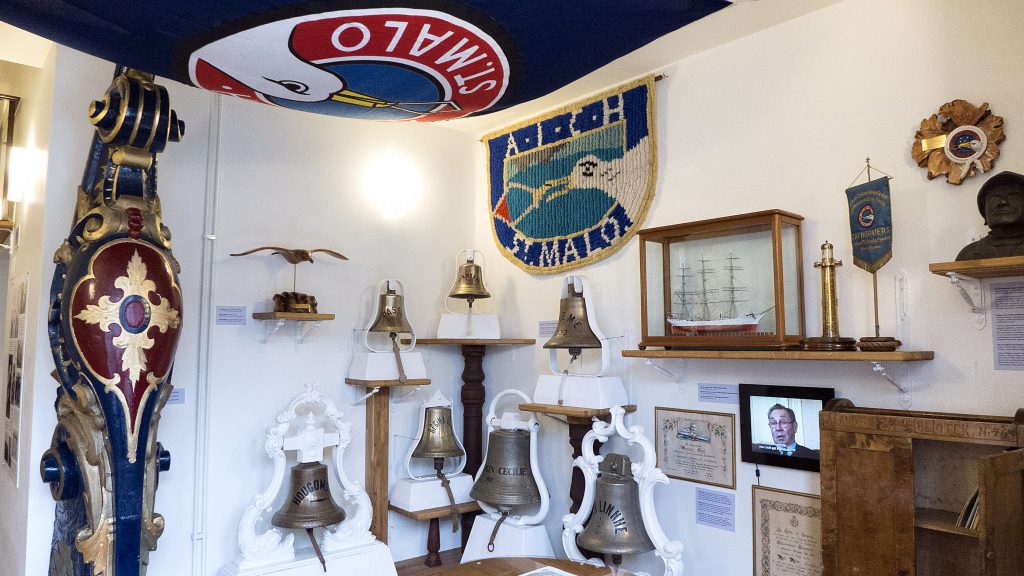 Ett museirum visar skeppsklockor, plaketter, nautiska memorabilia, ett modellfartyg, en byst, en vävd banderoll och en TV-skärm som visar en persons ansikte – som hyllar sjöfartshistoria och legender som de som trotsade Kap Horn.