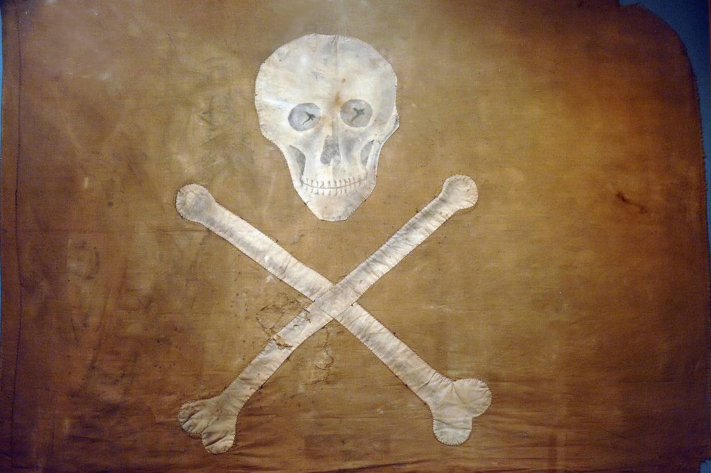 En väderbiten piratflagga från kuriosasakabinettet, med en vit dödskalle och korsben mot brun bakgrund.