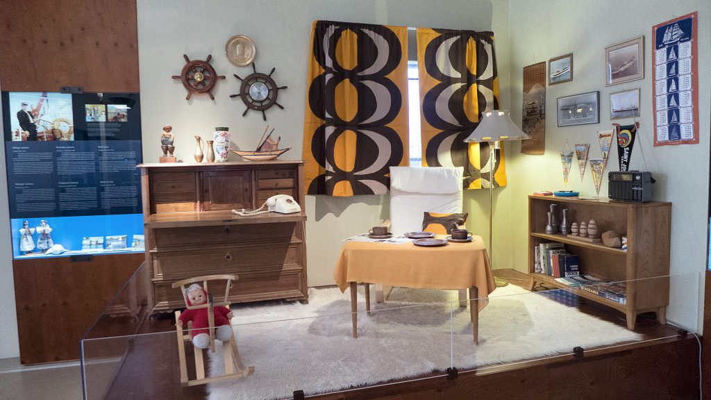 En vintage vardagsrumsmonter med nautisk inredning, ett matbord, trämöbler, en docka i en stol och orangemönstrade gardiner framkallar Mariehamns maritima charm, allt bakom en glasbarriär i denna museiutställning.