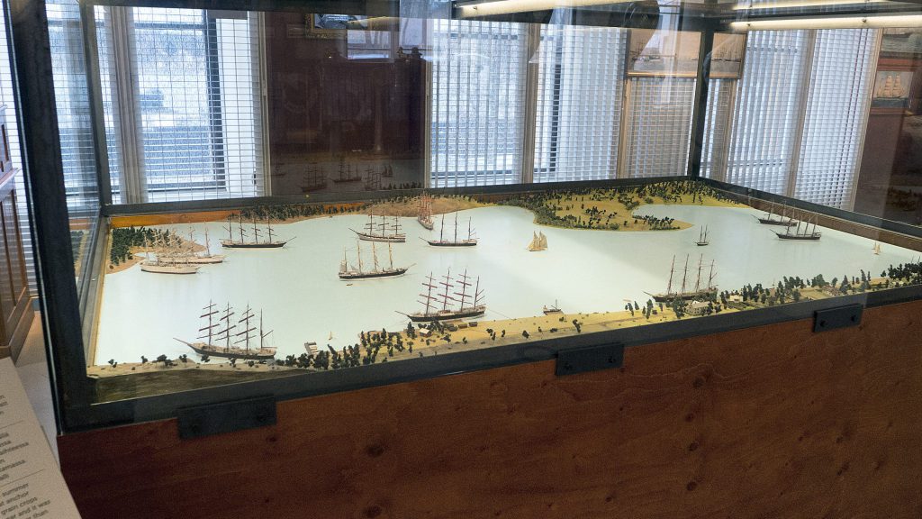 En glasmontre innehåller ett detaljerat diorama av en kustlandskap i Mariehamn, med flera segelfartyg, träd och en pittoresk strandlinje.