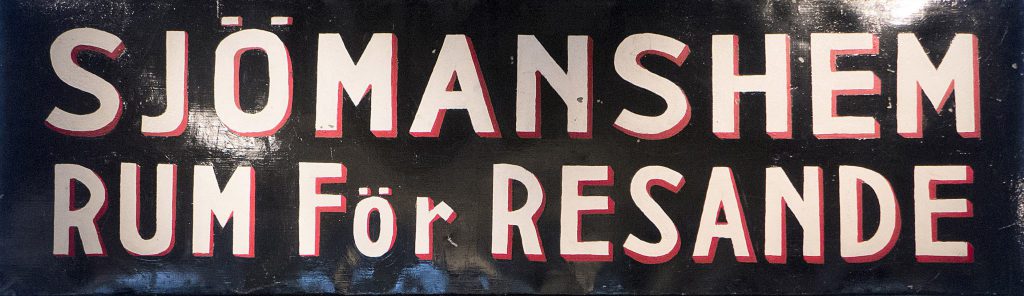 En svart skylt med vit och röd text där det står SJÖMANsHEM RUM FÖR RESANDE på svenska, sett i Mariehamn.