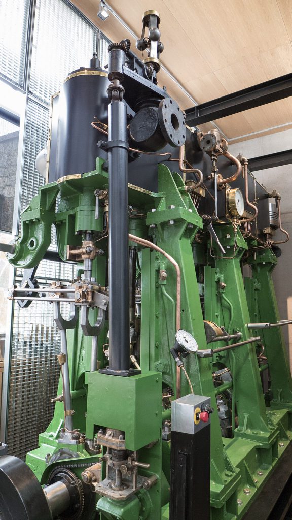 Stor grön och svart industriell ånglok med metallrör, mätare och kontrollknappar – ett imponerande exempel på Maskinsjöfarten – uppställd inomhus med naturligt ljus.