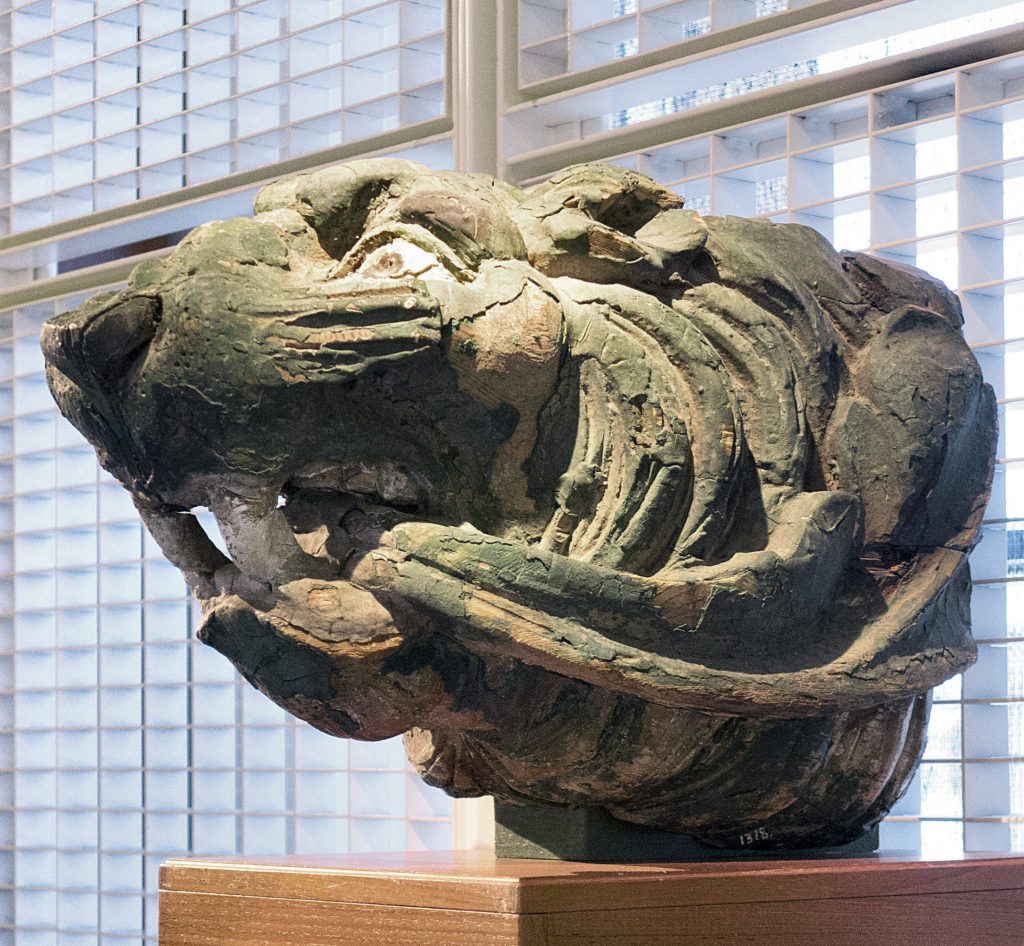 En väderbiten träskulptur av ett rytande tigerhuvud, inspirerad av Segelsjöfarten, visas på en piedestal framför persienner.