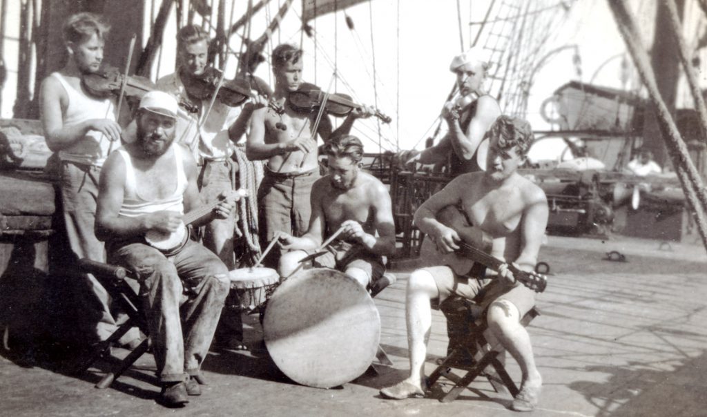 En grupp män på ett skeppsdäck, förkroppsligar Segelsjöfartens anda, spelar musikinstrument – inklusive fioler, gitarr, banjo och trummor – medan de sitter och bar överkropp i solljuset.