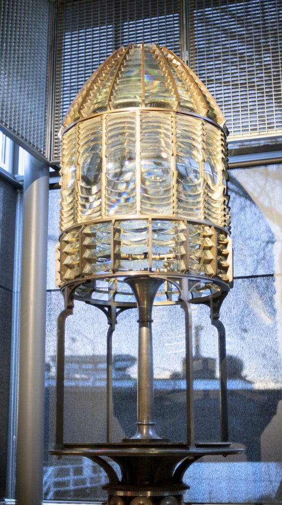 En stor Fresnel-lins, som vanligtvis används i fyrar för Sjösäkerhet, visas inomhus på ett metallstativ med en rutnätsliknande bakgrund.
