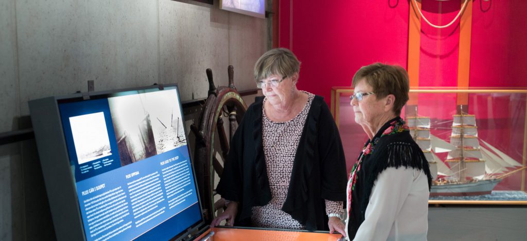 Två äldre kvinnor läser information på en bildskärm om sjösäkerhet på ett sjöfartsmuseum, med en fartygsmodell och ett fartygsratt synliga i bakgrunden.