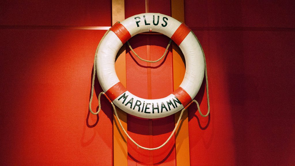 En vit flytväst med röda band och texten PLUS MARIEHAMN, som symboliserar Sjösäkerhet, hänger på en röd vägg.