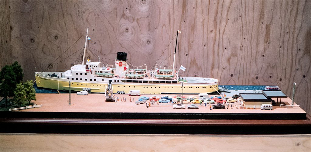 En detaljerad modell av Ålandsfärjan som ligger förtöjt i en miniatyrhamn, omgiven av parkerade bilar, små byggnader, träd och små figurer.
