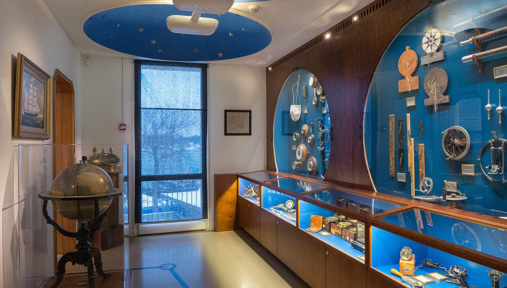 En museumsutställning visar navigationsinstrument och maritima artefakter i glasmontrar, med en jordglob och inramat konstverk synliga i rummet, som belyser navigationens utveckling genom historien.
