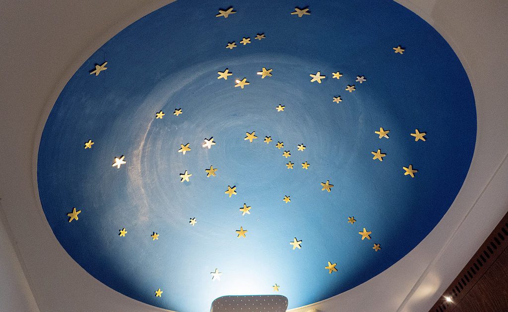 En blå cirkulär takmålning med gyllene stjärnformer utspridda över, som liknar en natthimmel eller ett stjärnbildsmönster, frammanar den tidlösa konsten att navigera med hjälp av stjärnljus.