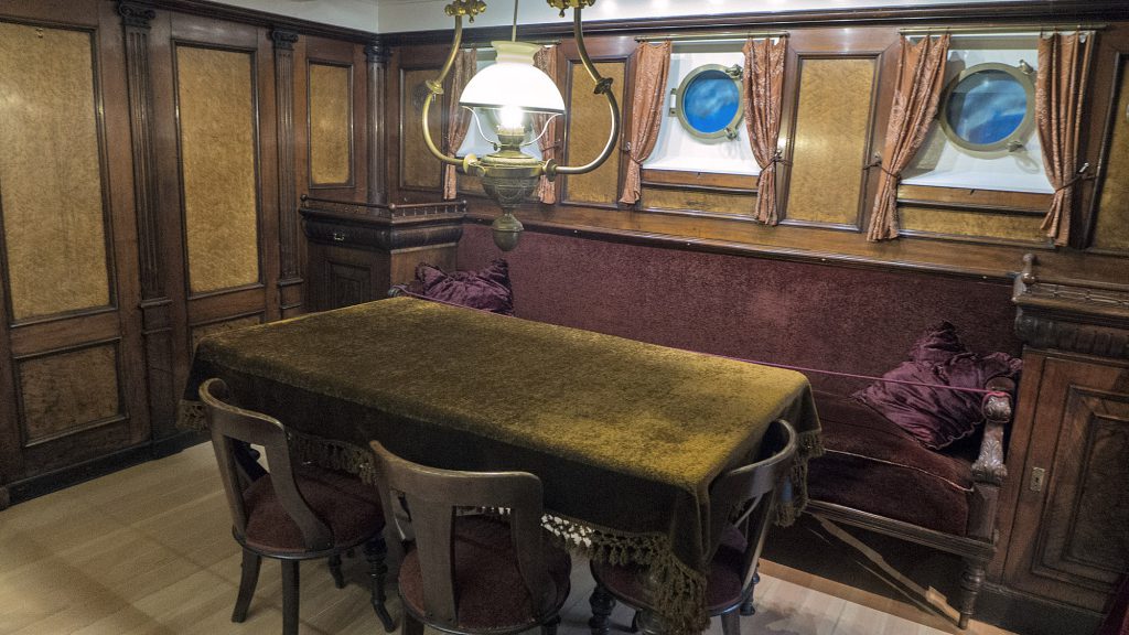 En matplats i en vintagekabin med träpaneler på väggarna, ett bord täckt med en brun duk, fyra stolar, en stoppad bänk och två runda fönster med ventiler.