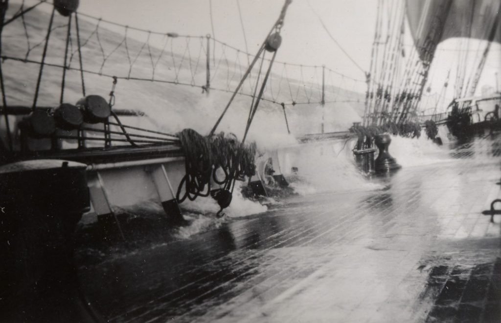 Black and white photo of a ship's deck tilted by rough waves, with water washing over the side and ropes and equipment visible.