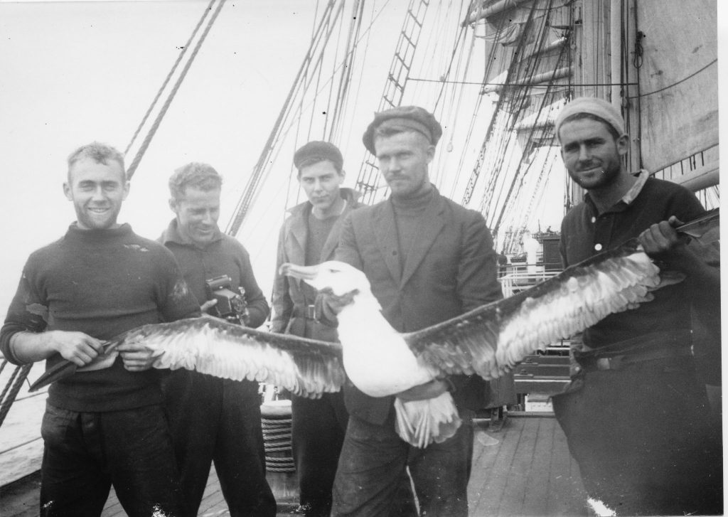 Five men stand on a ship's deck holding a large albatross with outstretched wings.