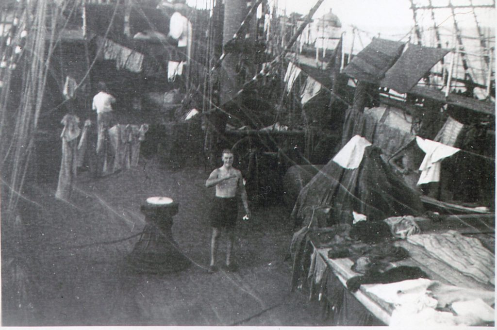 Black and white photo of a ship's deck with laundry hanging, linens laid out and a person standing in the middle looking into the camera.