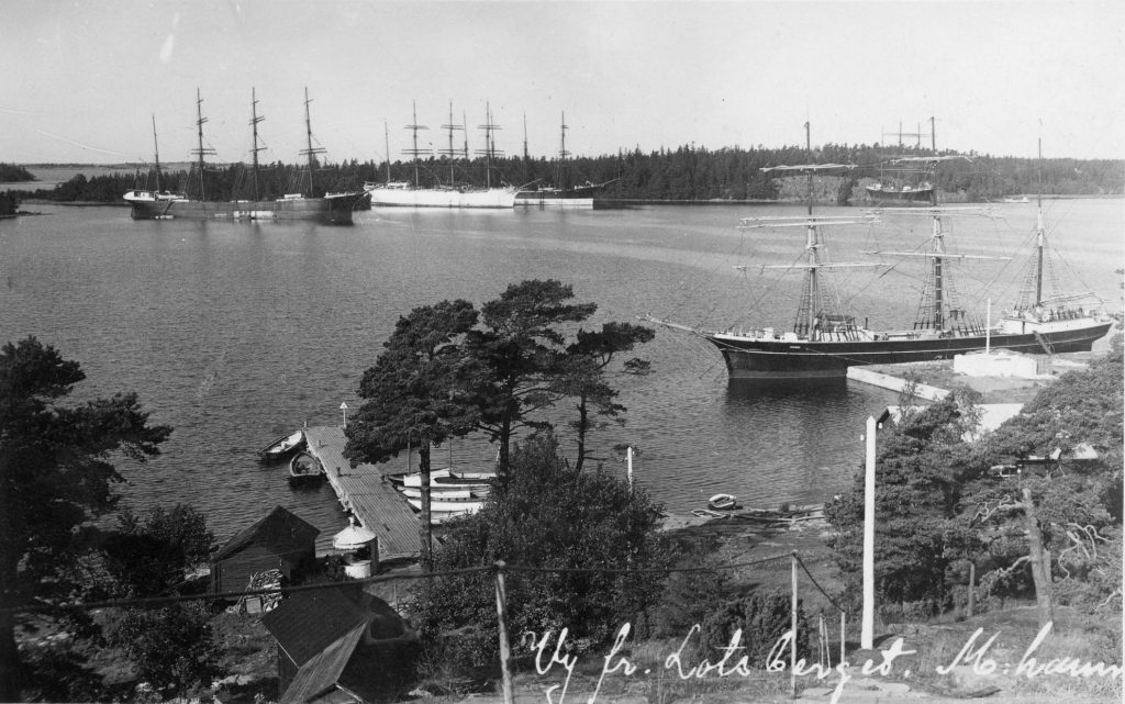 Black and white photo of several large sailing ships moored near Mariehamn's shoreline, with trees, small boats and wooden jetties in the foreground.