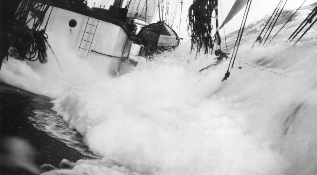 En stor våg slår över däcket på ett segelfartyg, lutar fartyget och skvätter vatten bland rep och utrustning – en dramatisk påminnelse om sjösäkerhetens betydelse till sjöss.