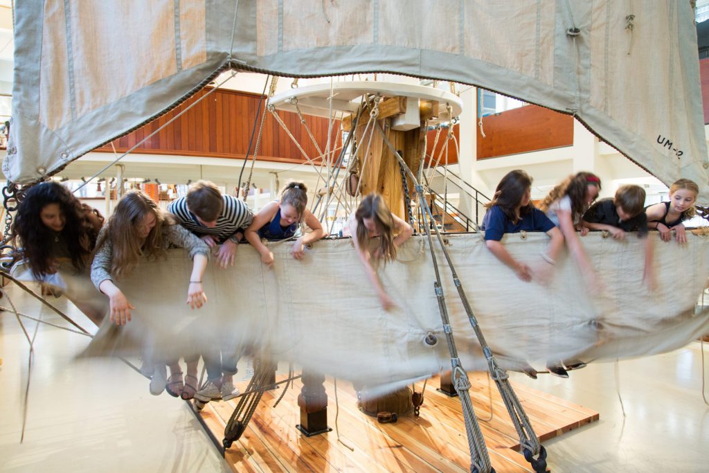 A group of children lean over the edge of a ship's deck inside a museum, reaching down and interacting with ropes and sails.