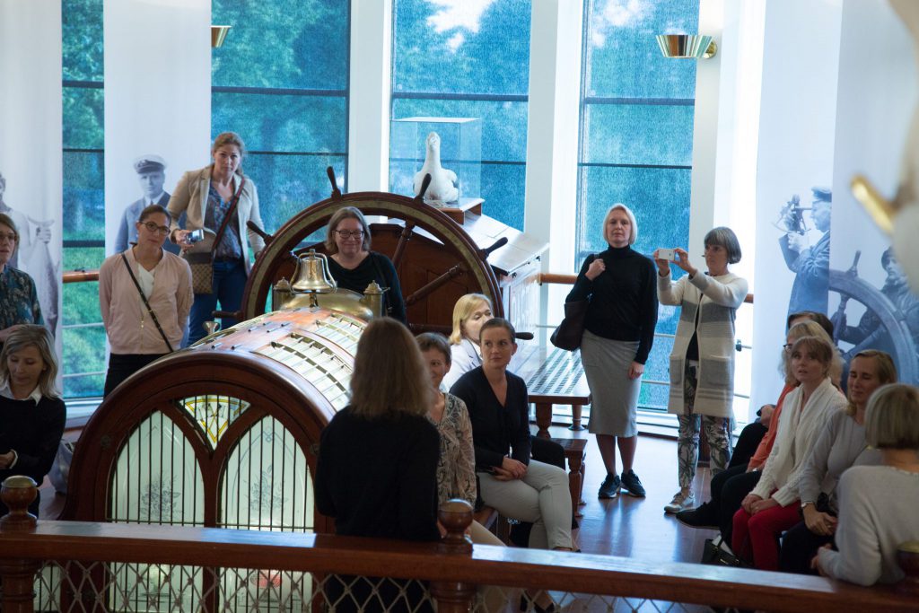 En grupp kvinnor, de flesta blonda, några gråhåriga eller rågblonda, står på en plats i museets byggnad som föreställande en imitation av ett poopdäck på en båt. Längs med väggarna hänger banners med svartvita fotografier av sjömän och tre fönster längst bak i rummet, där man ser dagsljus och grönska. På poopdäck finns roder och en inglasad, uppstoppad albatross finns på konsolen där rodret sitter fast. Längst fram i bilden löper en reling.