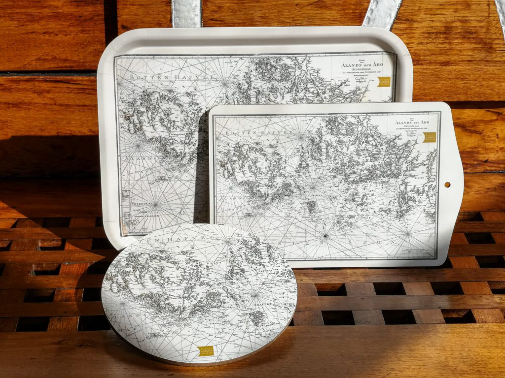 På en bakgrund av mellanmörkt trä, "golvet" med hål i rutmönster och "väggen" lite gulare trä med vita målade linjer högst upp finns tre ljusa föremål ställda på rad framifrån och bak. Längst fram till vänster av bildens mitt finns ett runt grytunderlägg it trä med sjökort som motiv. Bakom den längre mot höger av bilden syns ett tjockt skärbräde med samma motiv. I bildens centrala del längst bak i raden av föremål finns en bricka med samma motiv som de andra två föremålen.