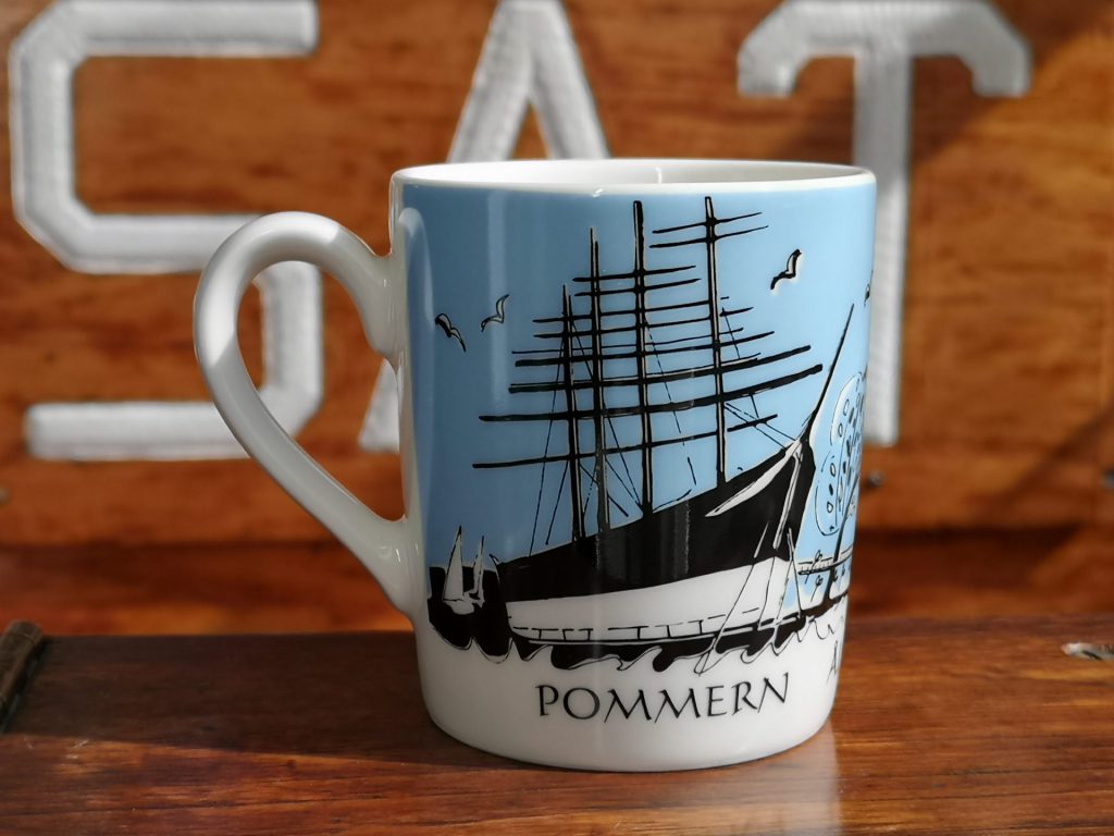En vit mugg med en svart skeppsillustration märkt POMMERN på blå bakgrund, placerad framför en träyta med delvis synliga vita bokstäver – perfekt som inspiration när du vill planera ditt besök.
