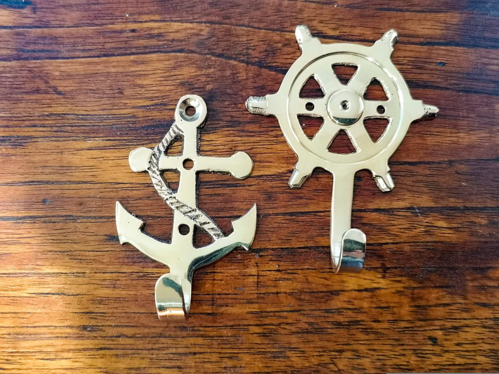 On a background of dark wood, two brass hooks are visible. To the right and just below the center of the picture is a hook representing an anchor, with the hook at the bottom, and a rope running diagonally across the anchor from its ring at the top. To the right and a little higher up in the picture is a brass hook representing a rudder, with a longer hook at the bottom.