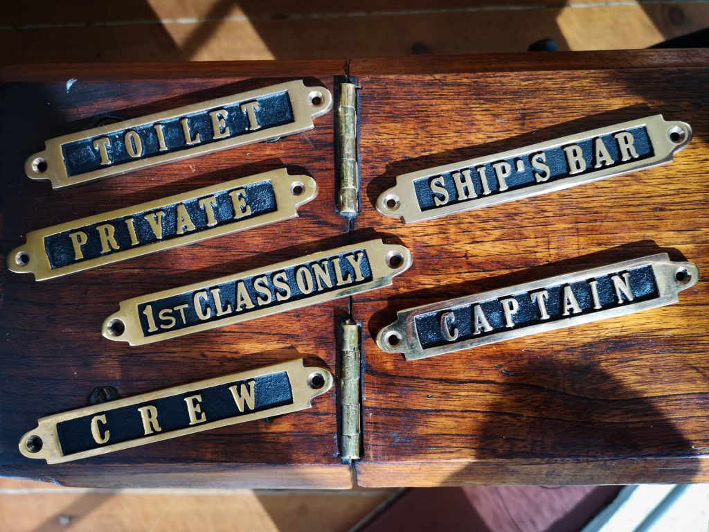 Museokaupan kuusi messinki- ja mustametallikylttiä lepää puupinnalla, kuhunkin kaiverrettu TOILET, PRIVATE, ONLY 1ST CLASS, CREW, SHIP'S BAR ja captain.