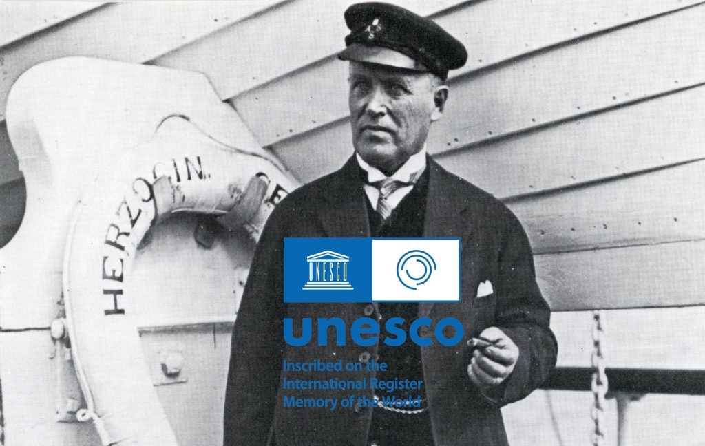 Svartvitt foto av Gustav Erikson i uniform som står på ett fartygsdäck, med en livboj märkt HERZOGIN; UNESCO-logotyp och textöverlägg längst ner.